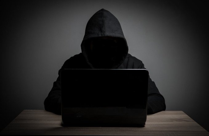A hooded figure sits in darkness behind a laptop, symbolizing the anonymous hacker who exploited BonkDAO's governance system to drain $20 million in $BONK tokens from the treasury after acquiring $4 million in voting power.