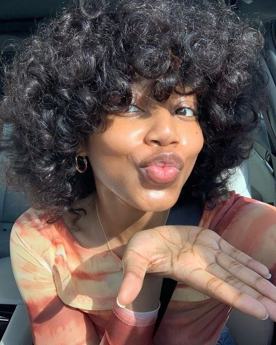 A woman with voluminous curly black hair wearing an orange and yellow tie-dye top sits in a car, puckering her lips playfully while gesturing with her hand.