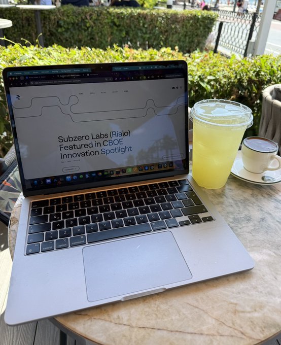 A MacBook Pro laptop sits on an outdoor cafe table displaying 'Subzero Labs (Rialo) Featured in CBOE Innovation Spotlight' next to a glass of lemonade and a cup of coffee.