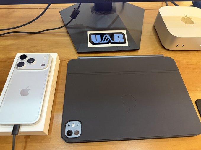 An iPhone 15 Pro and an iPad Pro rest on a wooden desk alongside a Mac Mini, with a "UAR" sticker visible on the display stand—showcasing the upgraded Apple gear the poster mentions while asking about their OneKey hardware wallet.