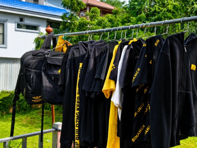 A clothes rack outdoors displaying black and yellow 'Binance' branded merchandise including t-shirts, hoodies, and a backpack.