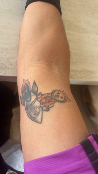 A close-up photograph of a person's forearm displaying a small tattoo of an astronaut riding a rocket, accented with orange and black ink.