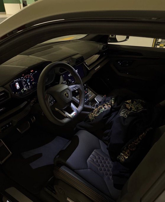 The interior of a luxury car, featuring a digital dashboard, a steering wheel with a Lamborghini logo, a gear selector, and a jacket resting on the passenger seat.