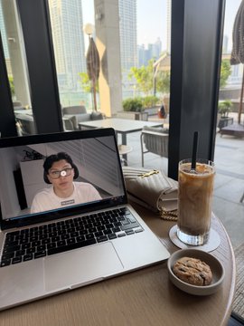 A laptop on a wooden table displays a video call with a woman wearing glasses, accompanied by an iced coffee drink and a cookie on a saucer, with a city skyline visible through the window.