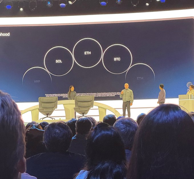 A panel discussion takes place on stage with a screen displaying cryptocurrency logos (SOL, ETH, BTC, XRP) at what appears to be a Robinhood ($HOOD) event, highlighting the platform's expansion into multiple asset classes including crypto trading as mentioned in the post.
