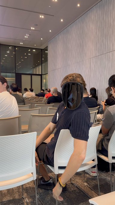 A person wearing a black Apple Vision Pro headset sits on a white chair in a conference or presentation setting with other attendees visible in the background.