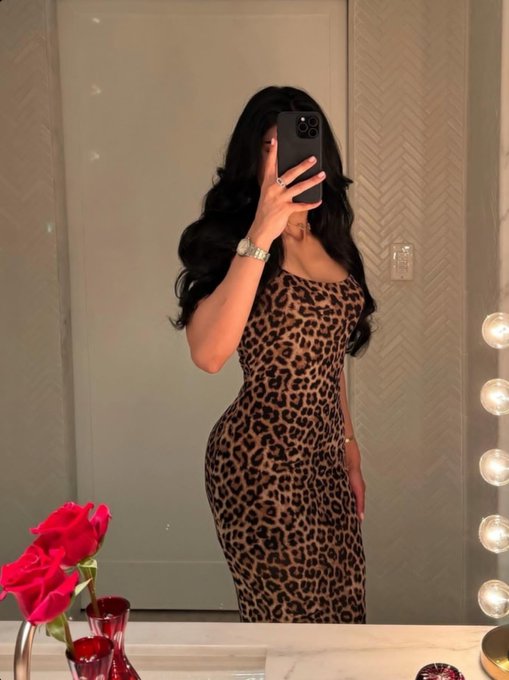 A woman with long dark wavy hair takes a mirror selfie in a leopard print dress, wearing a watch and rings, with pink roses visible in the foreground.