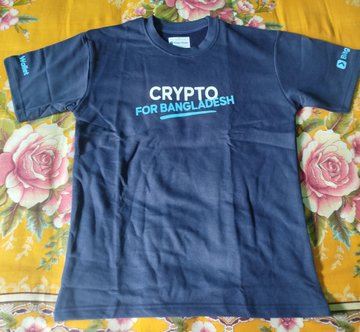 The image features a navy blue t-shirt laid out flat, with the text "Cypto for Bangladesh" written across the front of it. The shirt appears to be new and unused, as indicated by its pristine condition. The background is a simple yellow fabric, providing a contrasting backdrop that makes the black t-shirt stand out even more.