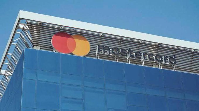 The image shows the exterior of a modern glass building featuring the Mastercard logo—two overlapping red and yellow circles—and the "mastercard" name in white lettering on its facade, visually representing the company that has just completed its acquisition of stablecoin infrastructure firm BVNK for $1.8 billion.