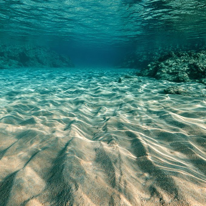 An underwater view showing a rippled sandy seabed illuminated by sunlight filtering through clear turquoise water.