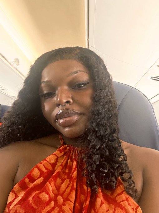 A close-up selfie of a woman with long curly hair wearing an orange and red patterned halter top, seated inside an airplane.