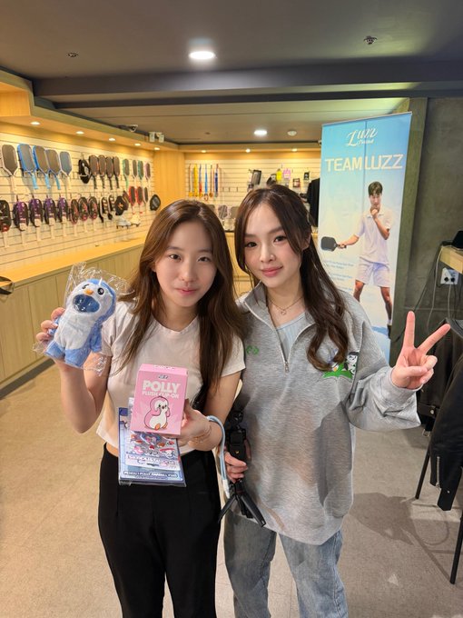 Two young women posing together inside a Luzz pickleball shop, with the left woman holding a blue plush toy and a pink 'POLLY MOLA' package, while a 'TEAM LUZZ' banner hangs in the background.