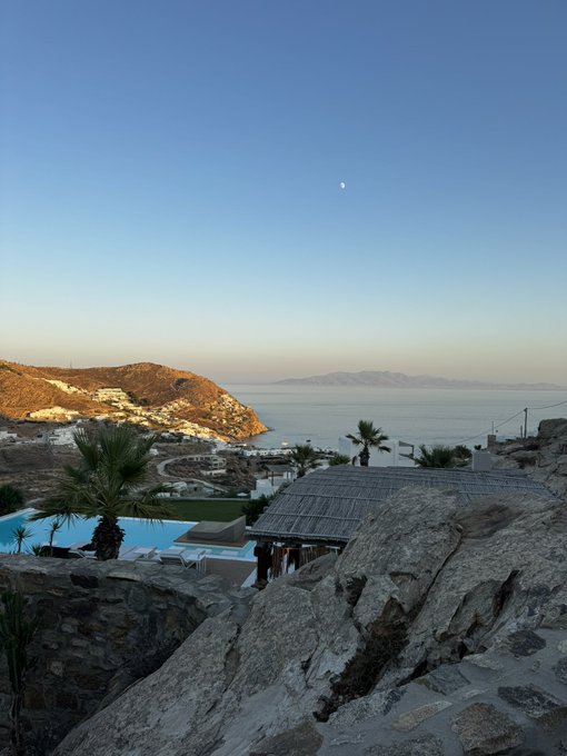 A stunning sunset view from a clifftop resort overlooking a Mediterranean bay, a distant island, and a moon rising in the twilight sky — the perfect "Bender activation" backdrop.