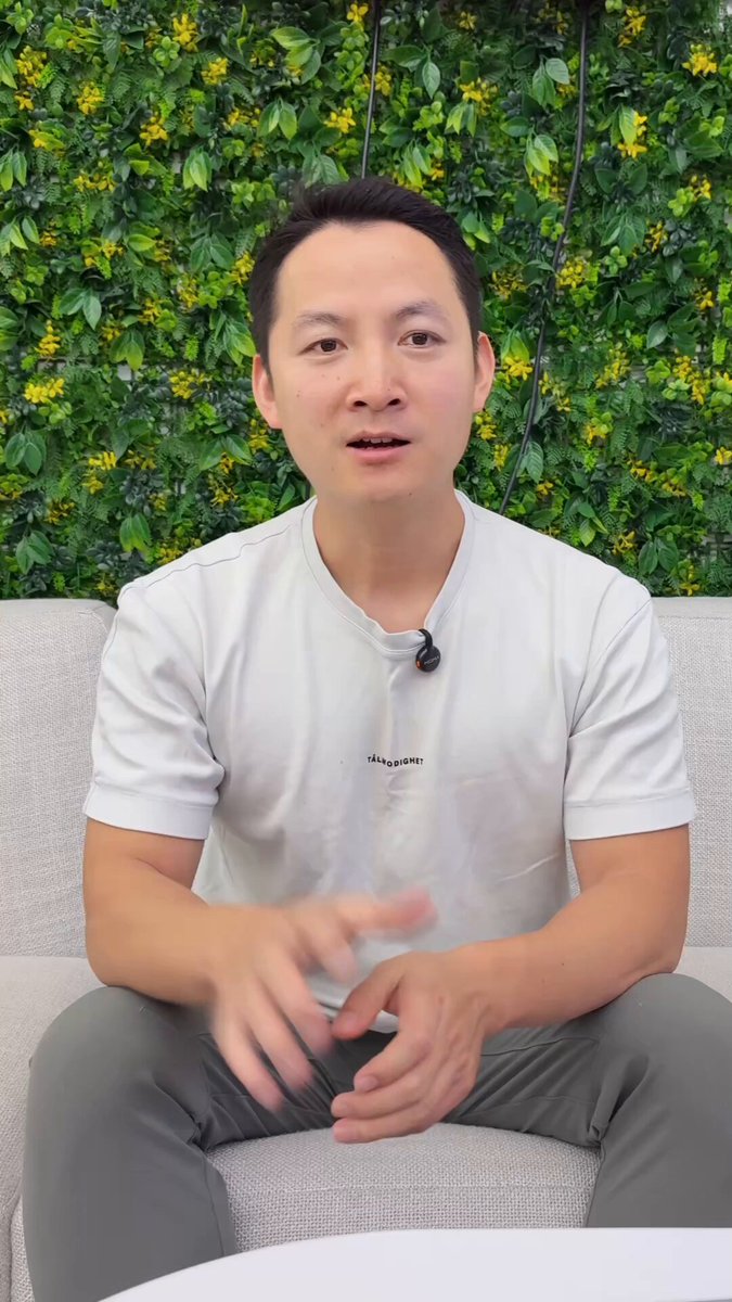The image features a man sitting on a couch, wearing a white shirt with black text. The man appears to be talking or posing for a photo, as he has his hands together while looking at the camera. The background of the image consists of a green wall and a plant, creating an interesting visual contrast against the man's attire. The post includes a caption that reads "OP_CAT Layer 为什么值得关注？ 因为比特币的可编程性和扩展性仍然存在很大的限制". This caption suggests that the image may be related to a discussion or conversation about OpCats, which are digital assets that can be bought and sold on the secondary market.