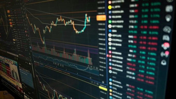 A close-up photograph of a trading screen displaying Bitcoin price charts with candlestick patterns, volume bars, and a list of cryptocurrency prices showing red and green values.