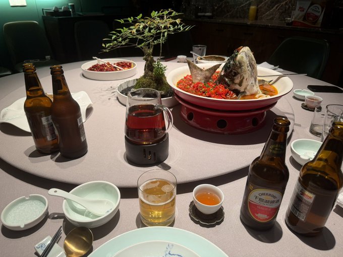 The image shows a dining table with various items, including several bottles of beer, bowls, cups, spoons, and a potted plant. The scene appears to be set for a meal or gathering at someone's home. The presence of multiple bottles suggests that there might be drinks being served during the event. The variety of dishes on the table indicates that it could be a social gathering where people can enjoy food and beverages together.