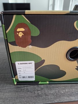 A cardboard shoebox with a green and tan camouflage pattern featuring the iconic BAPE ape head logo and a white product label.