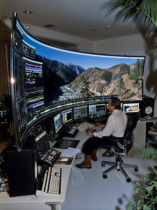 A man in a white shirt sits at an elaborate curved multi-screen workstation displaying editing software and mountain scenery, working at what appears to be a high-end post-production setup that humorously contrasts with the modest $180 portfolio referenced in the post.
