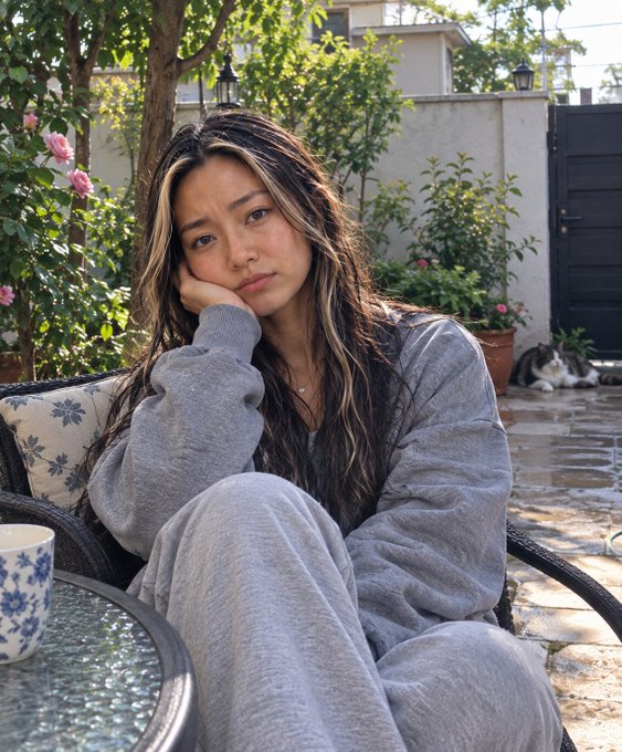 A young woman with long highlighted hair wearing a gray sweater and sweatpants sits looking bored with her chin resting on her hand in an outdoor patio setting, with a blue and white teacup on a table and a cat resting in the background.