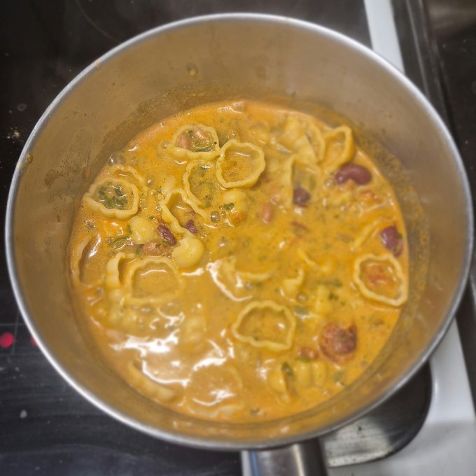 A metal pot on a stovetop contains a creamy orange soup with tortellini pasta, red beans, and green herbs.