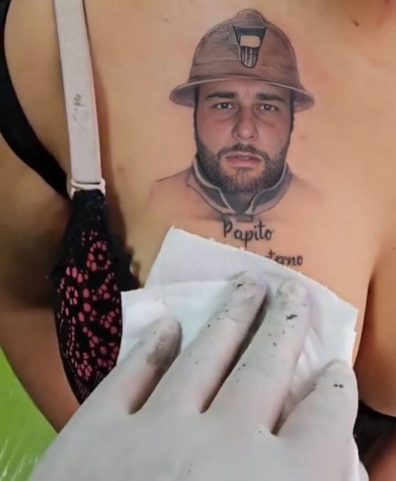 A close-up of a woman's chest featuring a tattoo of a bearded man wearing a mining helmet above the words 'Papito' and 'terno', while a soot-stained hand holds a white tissue nearby.