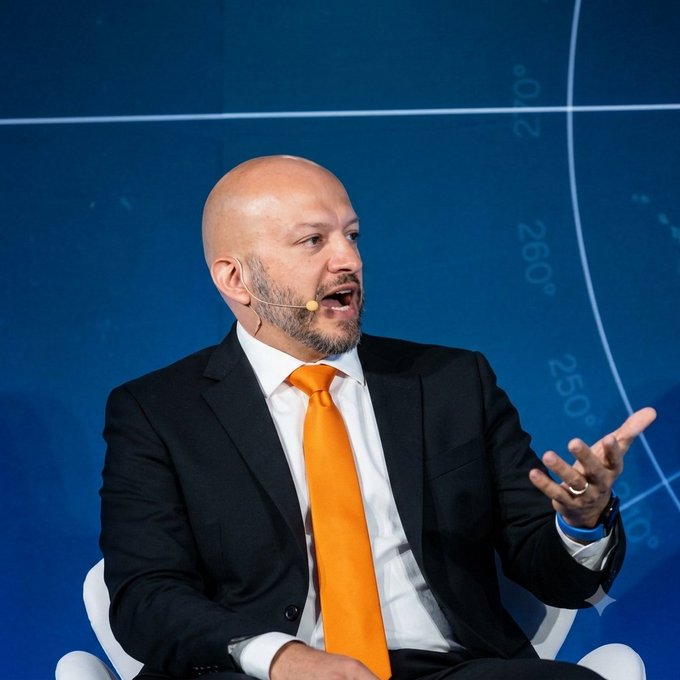 A bald man with a beard in a dark suit and orange tie speaks while gesturing, appearing to present at a conference with a blue background displaying degree markings.