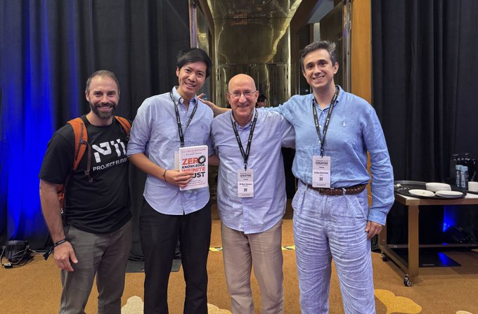 Four men pose together at an event; one holds a book titled 'ZERO TO ONE,' and another wears a shirt reading 'PROJECT FIRST.'