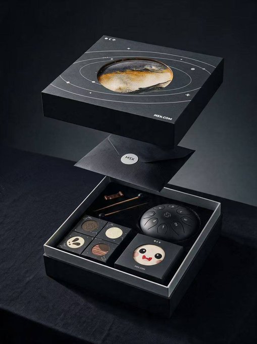 A black NSX gift box featuring an image of a moon on the lid, with the lid lifted to reveal an assortment of chocolates, including planets, stars, a panda design, and decorative chocolate pieces.