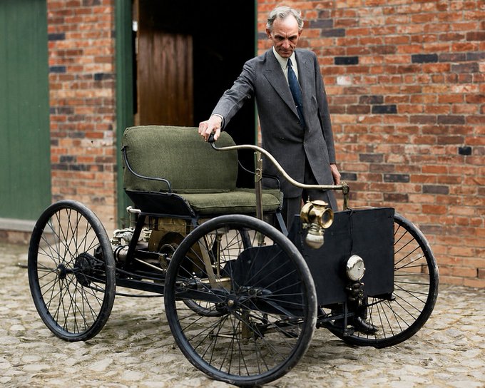 Photograph of an older man in a grey suit standing behind a vintage four-wheeled horseless carriage on a cobblestone street in front of a brick building.