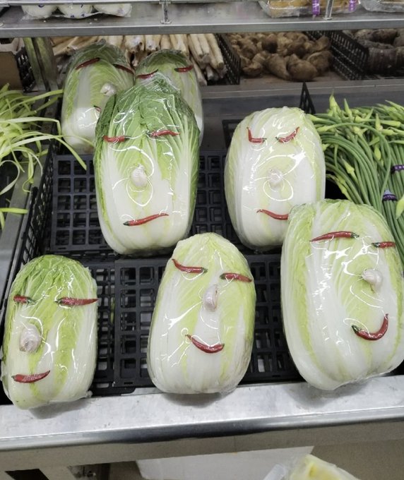 Napa cabbages with faces drawn on them are displayed in plastic wrap on a supermarket shelf, surrounded by green beans and ginger. The smiling, relaxed faces of the unassuming cabbages serve as a visual metaphor for the post's message: these veggies look calm and content because nobody is paying attention to them yet, just like how the most profitable opportunities exist quietly before the crowd notices and drives prices up.