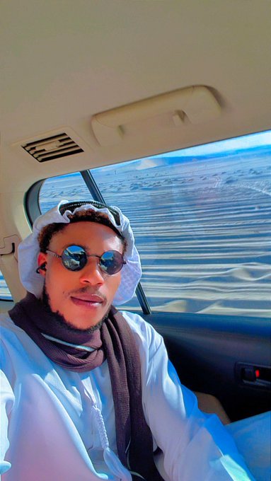 A man wearing sunglasses and a traditional white thobe with a headscarf and dark scarf takes a selfie from inside a vehicle, with rippled desert sand dunes visible through the window behind him. The setting suggests a desert location that aligns with the post's announcement about an upcoming giveaway winner.