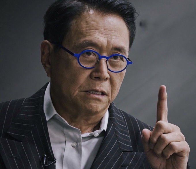 An elderly man wearing blue-rimmed glasses and a pinstripe suit gestures upward with one finger, his expression intense and cautionary. The accompanying text attributes a dire financial warning to Robert Kiyosaki, who predicts "the biggest crash in history" driven by the "AI frenzy, War in Iran, too much debt and the retiring baby boomer generation," advising followers to buy gold, silver, and bitcoin. The man's serious demeanor and emphatic pointing finger visually reinforce the urgency and gravity of Kiyosaki's dire market predictions.