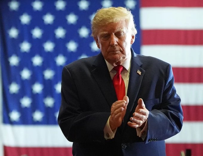 Donald Trump in a dark suit and red tie clapping with his eyes closed, standing in front of an American flag.