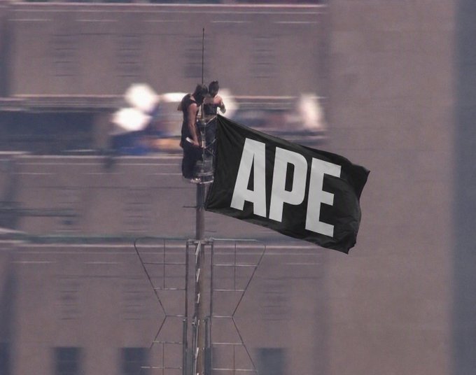 A person perched atop a pole raises a black flag with "APE" emblazoned in large white letters against the backdrop of a tall building, capturing the spirit of the accompanying post about embracing the $ape meme and rallying behind the playful "ape" phenomenon.