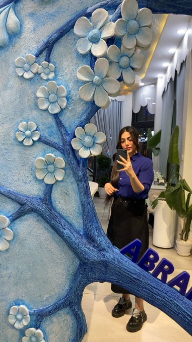 A woman taking a mirror selfie in what appears to be a hotel lobby, with a decorative blue floral sculpture in the foreground and partial text 'ABRA' visible.