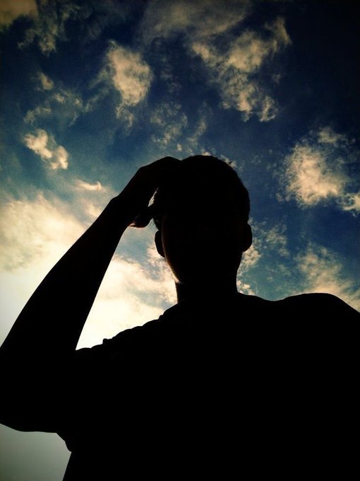 The image shows a silhouette of a person with their head tilted upwards, looking up at the sky. They are wearing a black shirt against a blue sky filled with clouds. The photo appears to be taken during sunset or sunrise, as suggested by the warm colors in the background. The caption "Gn CT The grind I still on The night is for the watchers" seems to be related to the image's content and could possibly be a reference to a popular YouTuber or a social media post about appreciating nature or enjoying outdoor activities during dusk or dawn hours.