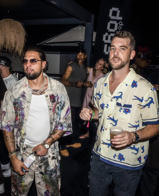 Two men pose at a nighttime social event; one wears a paint-splattered shirt and sunglasses while holding a drink, and the other wears a cream-colored shirt with blue lizard patterns.