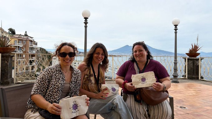 Three women sit on a terrace with a view of Mount Vesuvius and the Bay of Naples, each holding a small white canvas bag with a purple compass rose logo that reads "My Angel."
