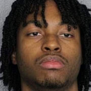 Close-up portrait photo of a young man with dark twisted dreadlocks, a light mustache and beard, photographed against a plain gray background in a style resembling a mugshot.
