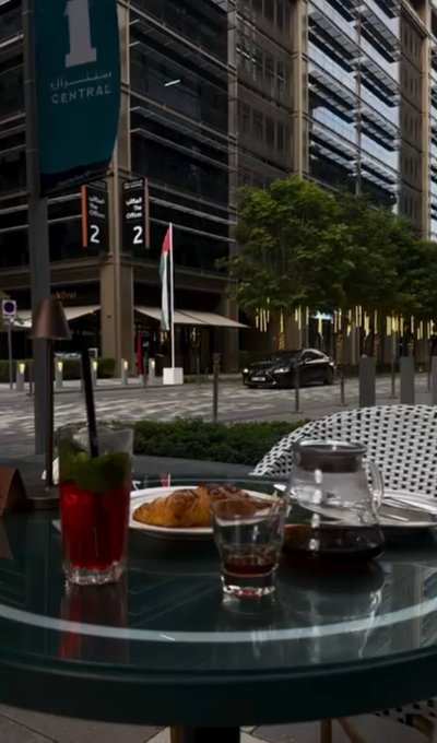 A glass table holds a red beverage, a glass of coffee, and food outside a building marked '1 CENTRAL', with modern glass architecture in the background.