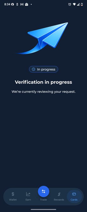 A mobile app screenshot showing a paper airplane logo with the text "In progress" and "Verification in progress. We're currently reviewing your request." above a bottom navigation bar with Wallet, Earn, Trade, Rewards, and Cards icons.