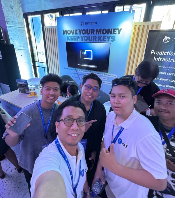A group of men wearing blue lanyards pose for a selfie in front of a Tangem booth advertising 'MOVE YOUR MONEY KEEP YOUR KEYS' at what appears to be a crypto conference.