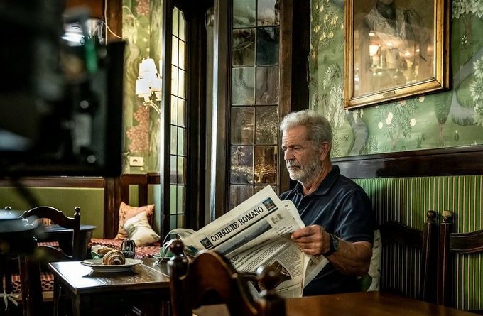 An older man with a grey beard sits in a pub reading a newspaper titled 'CORRIERE ROMANO', with a professional camera visible in the foreground.