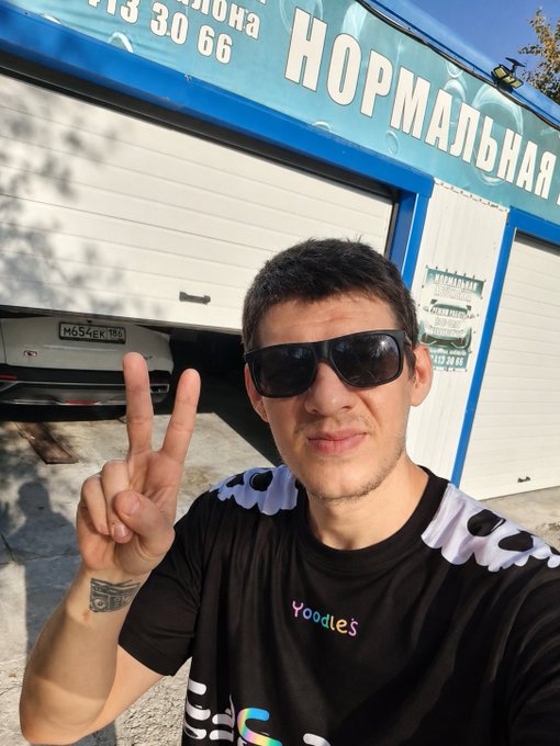 A man wearing sunglasses and a black 'Yoodles' t-shirt makes a peace sign in front of a building with Cyrillic signage reading 'НОРМАЛЬНАЯ' (Normalnaya) and a partially visible license plate 'M654EK 154'.