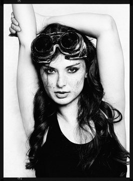 A black-and-white portrait of a young woman wearing steampunk-style goggles on her forehead, with arms raised behind her head and small decorative markings on her cheeks.