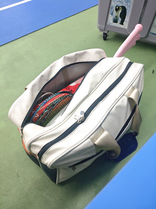 An open white rolling suitcase on a green sports court floor, containing a tennis racket and gear, with a pink tennis ball retriever sticking out.