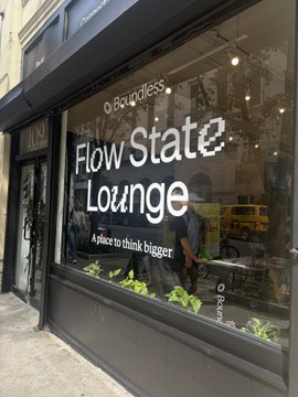 A street-level view of a storefront window with large white lettering reading "Boundless Flow State Lounge - A place to think bigger."