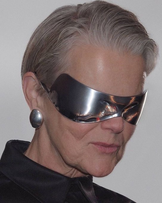 A close-up portrait of an older woman with gray hair wearing large, futuristic silver visor sunglasses and a black top.
