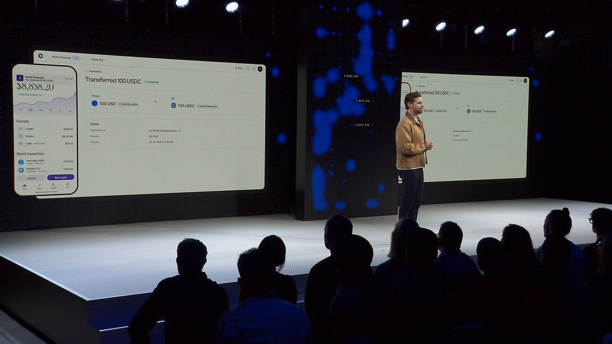 The image captures a man standing at an event, talking into a microphone while people sit behind him looking intently at two large screens displaying financial information. The screens are placed side by side on both sides of the room, allowing for clear visibility from all angles. A caption below the screen reads "Since we launched x402 last year, we've seen: → Over $100M in transactions → ?". It seems like an important discussion taking place about the performance of a specific cryptocurrency or the evolution of the industry itself.