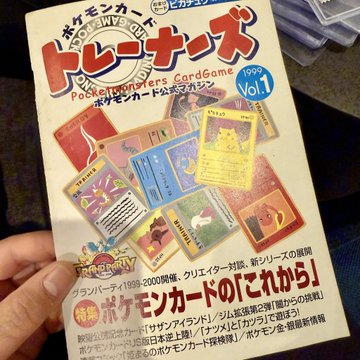 A hand holds a Japanese Pokémon Carddass 'Trainers' magazine from 1999, Volume 1, featuring various Pokémon cards on the cover.